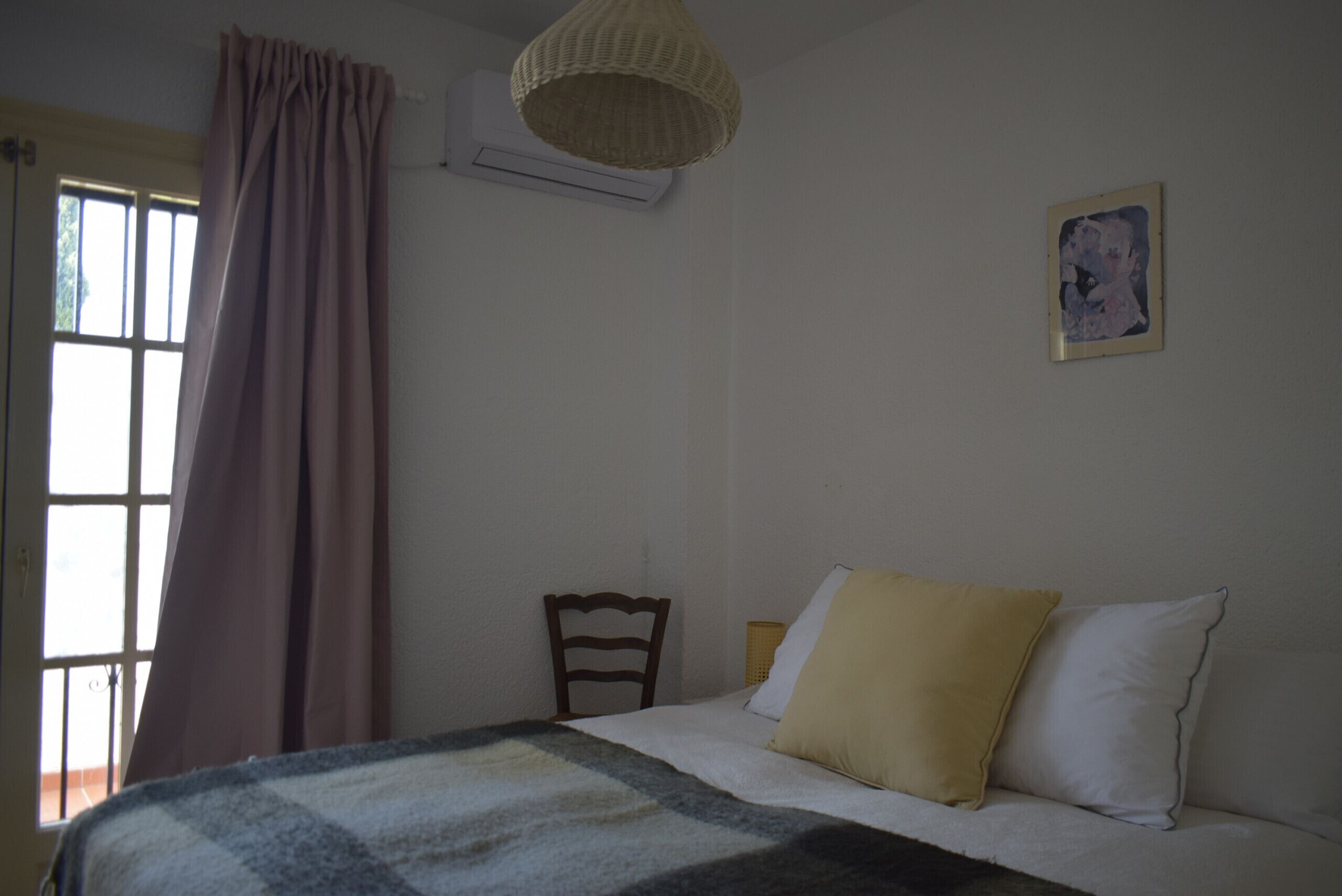 Une chambre à coucher comprenant un lit, une chaise, une fenêtre avec des rideaux, un appareil de climatisation et un luminaire fixé au mur.