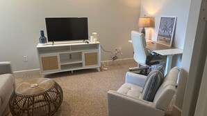 A living room with a flat-screen TV on a white entertainment unit, a wicker coffee table, a grey armchair, and a lamp on a side table. - Gorgeous 2nd Floor Unit in The Quarry, Minutes to downtown Naples and Mercato. (Naples)
