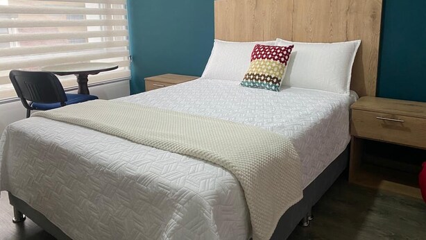 Uma cama bem arrumada com colcha branca, cabeceira de madeira e travesseiro colorido. Há uma cadeira e uma mesa pequena ao lado da cama.