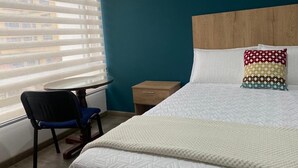 Quarto de hotel com cama, uma cadeira, uma mesa pequena e janela com persianas.
