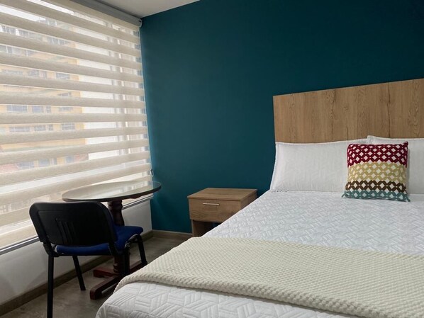 A hotel room with a bed, a chair, a small table, and a window with blinds.