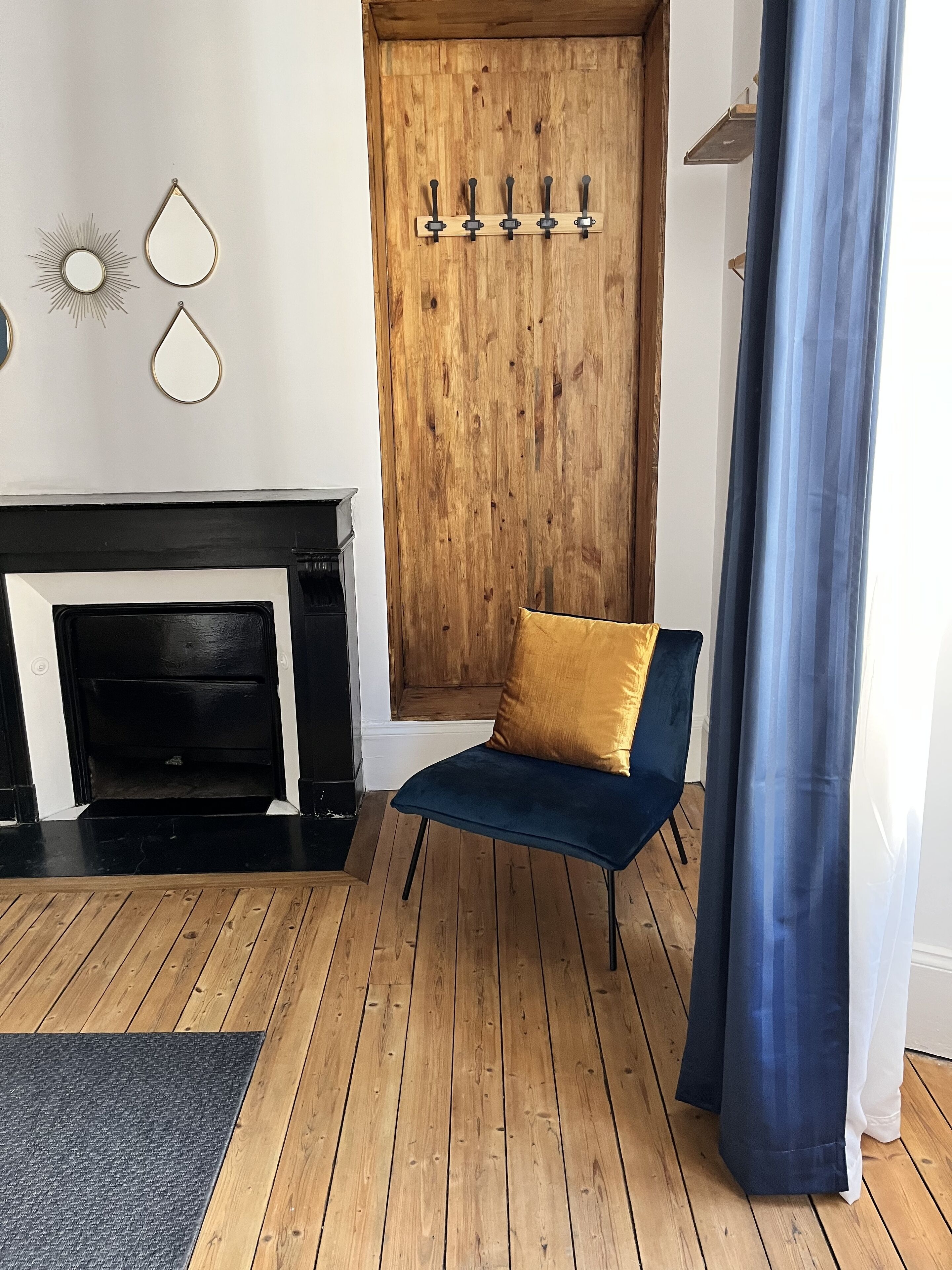 A room with a wooden floor, a blue chair with a mustard cushion, a wooden door, and a black fireplace.