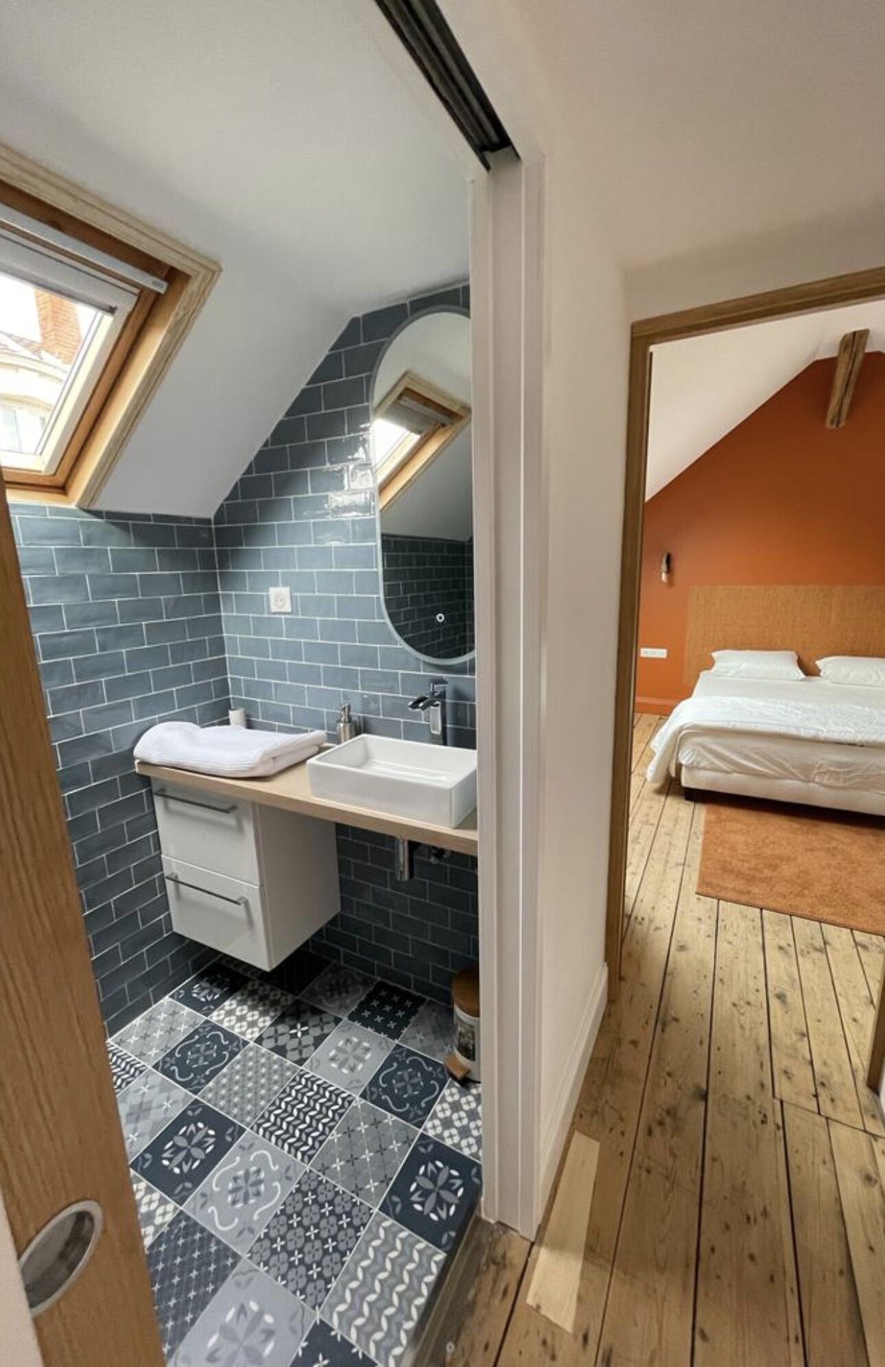A compact bedroom with a sloped ceiling, a small bathroom with a sink and mirror, and a patterned floor.