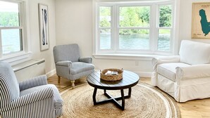 A room with a large window, a white sofa, a striped armchair, and a small table with a basket. - Lakefront Home on Lake Iroquois (Hinesburg)