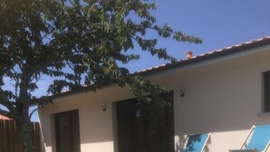 A wooden deck with a swimming pool, two lounge chairs, a tree, and a building with a tiled roof. - Maison Climatisée Avec Piscine Entre Montalivet et Soulac (Grayan-et-l'Hôpital)