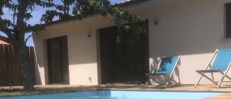 A wooden deck with a swimming pool, two lounge chairs, a tree, and a building with a tiled roof.
