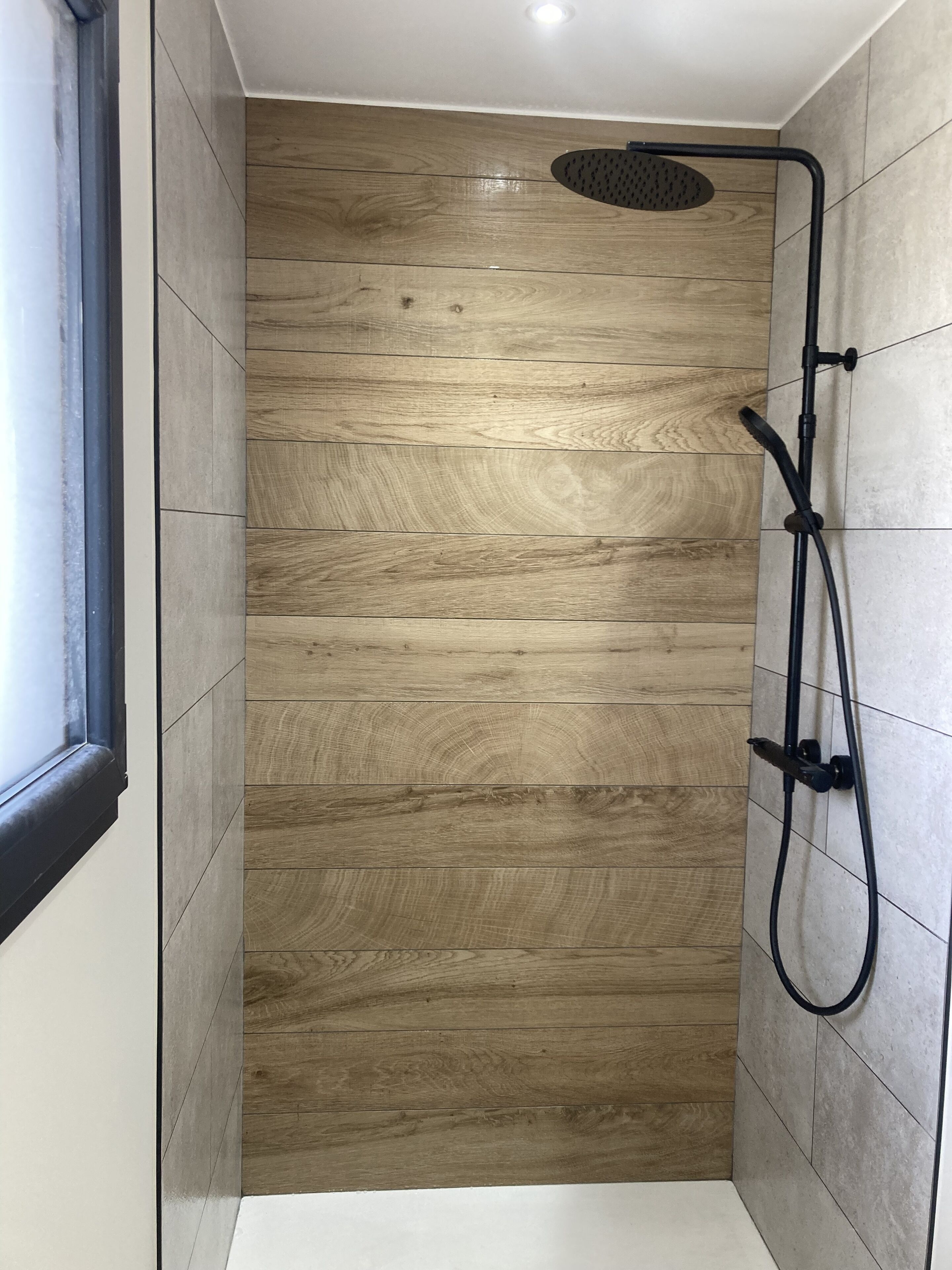 A shower with a wooden panel wall, a window, and a ceiling light.