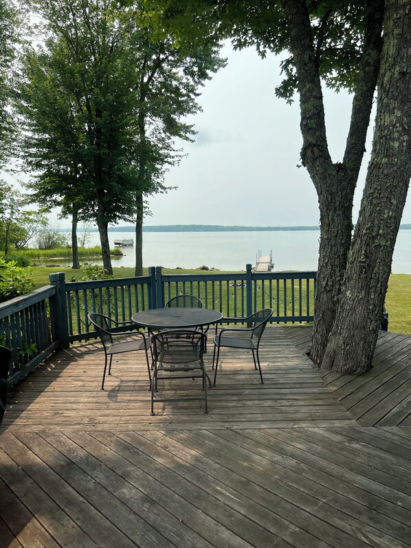 A wooden deck with a table and chairs, overlooking a calm lake with a distant shoreline.