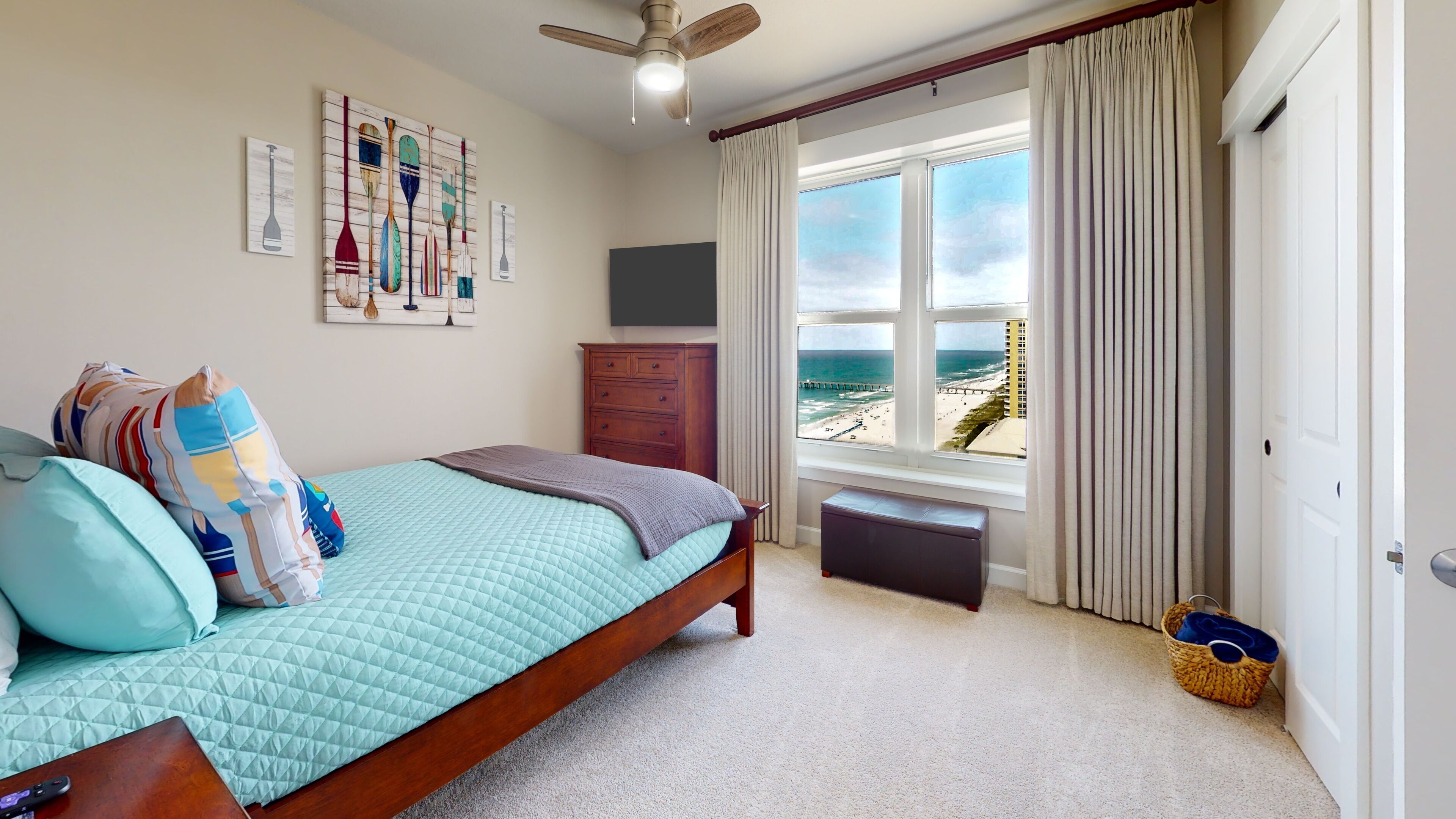A bedroom with a bed, a dresser, a window with a view of the beach, and a ceiling fan.