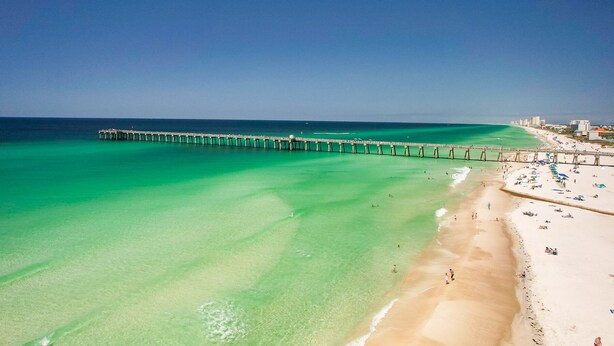 A beach with clear turquoise water, a pier extending into the sea, and a sandy shore with beach umbrellas and people. - Million dollar views, sleeps 10! Beachfront corner unit @ Grand Panama Resort! (Panama City Beach)