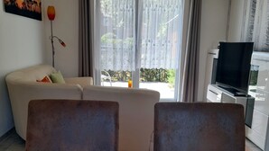 A living room with a sofa, two chairs, a dining table set for two, a TV, and a large window with curtains.