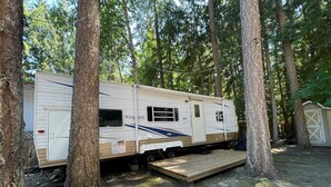 A Kingport RV parked in a wooded area with a wooden deck and a covered storage area. - Cozy cabin w/ Trailer, Sleeps 6 - 10 guests, Walking Distance to Beach, Hot Tub! (Swansea Point)