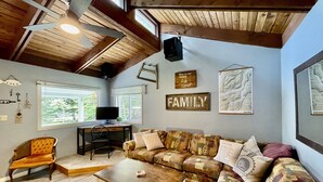 A cozy living room with a wooden ceiling, a large window, a sofa, a coffee table, and a TV. - Cozy cabin w/ Trailer, Sleeps 6 - 10 guests, Walking Distance to Beach, Hot Tub! (Swansea Point)