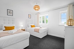 A modern hotel room with two beds, a window with curtains, a bedside table with a lamp, and a decorative basket. - 2 Bedroom House - Parking - Garden - Oldbury (Oldbury)