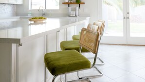 A modern kitchen with a white countertop, green cushioned bar stools, and a view of the outdoors through a glass door. - ༄The Agave Ranch༄ FREE Pool Heating & Arcade Room! (Phoenix)