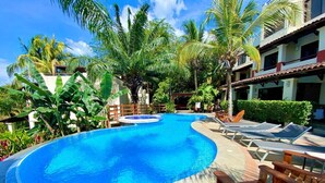 A swimming pool with lounge chairs, surrounded by palm trees and a building with a tiled roof.