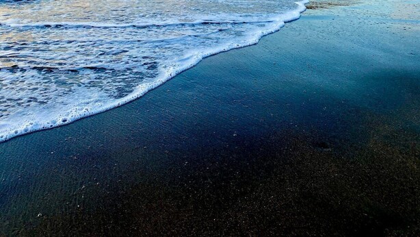 Une plage avec des vagues, un voilier et, au loin, une île couverte de forêts.