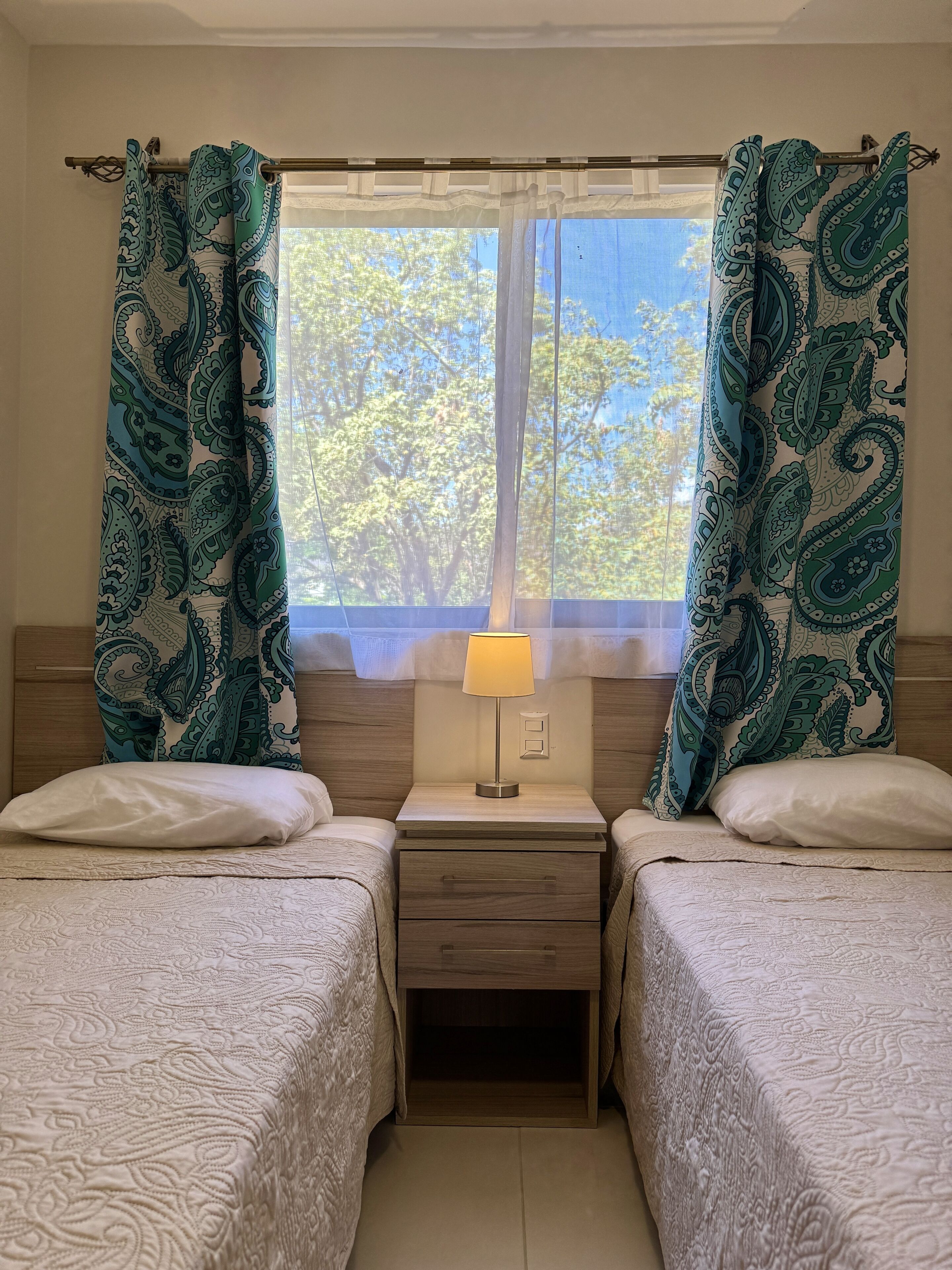 Une chambre avec deux lits, une table de chevet avec une lampe et une fenêtre avec des rideaux.