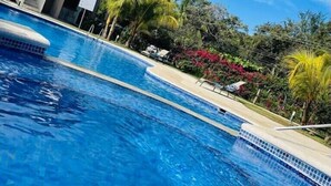 A swimming pool with clear blue water, surrounded by tropical plants and a modern building with balconies. - Parakeet Paradise 2nd floor Condo with Rooftop (Playas Del Coco)