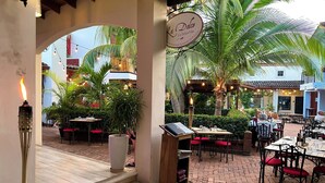Un área de comedor al aire libre con mesas y sillas, un cartel que dice 'La Dolce Vita International Restaurant & Pizzeria', y una escalera que lleva a un nivel superior.
