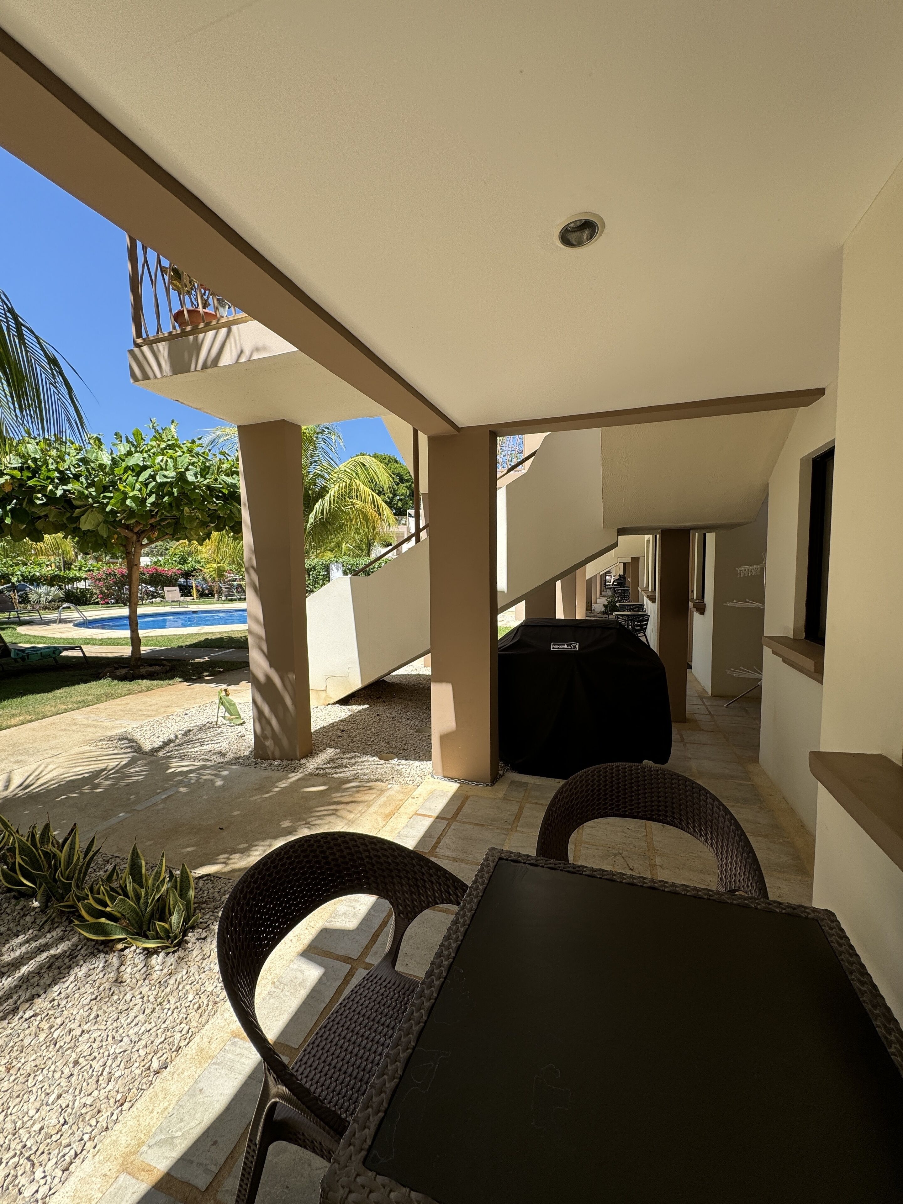 An outdoor patio with a table and chairs, a barbecue grill, and a pool in the background.