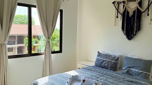 A neatly made bed with a patterned bedspread, a window with curtains, a wall-mounted air conditioner, and a macrame wall hanging.