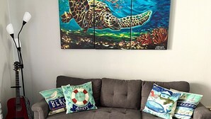 A living room with a grey sofa, colorful pillows, a guitar, and a painting of a sea turtle. - Sea Bottom Ground Condo (Playas Del Coco)