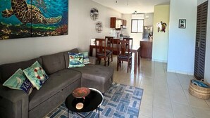 A living room with a gray sofa, a coffee table, and a painting of a sea turtle. There is a dining area with wooden furniture and a ceiling fan. - Sea Bottom Ground Condo (Playas Del Coco)