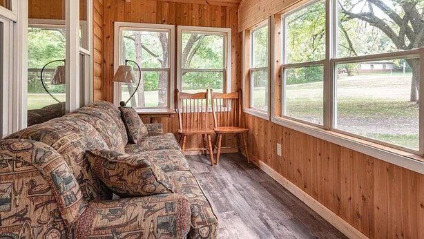 Interior acogedor de una cabaña con techo de madera, ventanas amplias, un sofá con cojines estampados y una mesa con sillas.