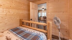 Habitación de cabaña de madera con una cama, un ventilador y una puerta que da a una zona de cocina.