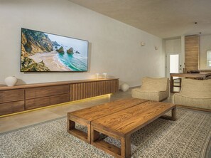 A modern living room with a wooden coffee table, a large flat-screen TV mounted on the wall, a woven sofa, and a wooden console table with decorative vases.