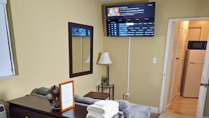 A hotel room with a dark wooden dresser, a flat-screen TV mounted on the wall, a small table with a lamp, and a gray sofa. - TheVacaVacationHomes (Lauderdale Lakes)