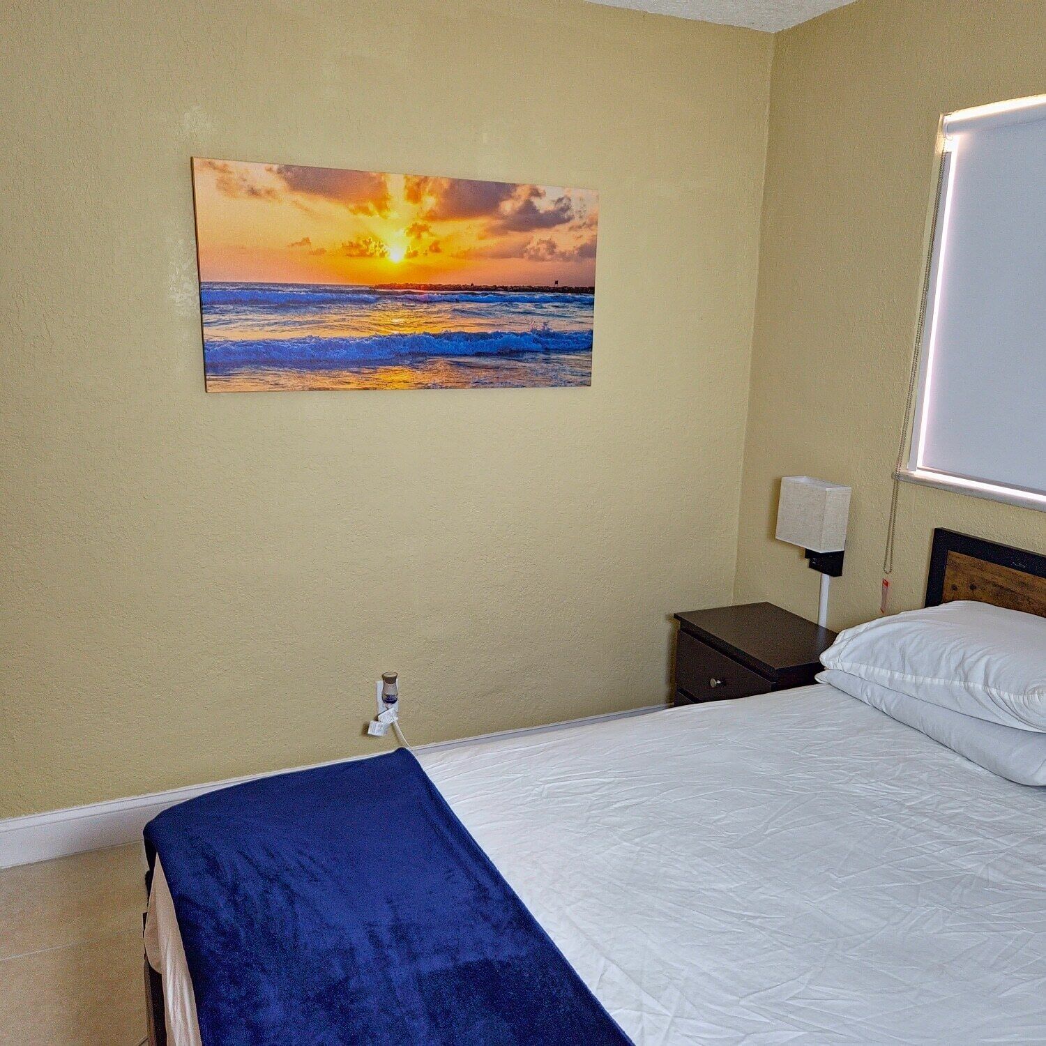 A hotel room with a bed, a nightstand, a lamp, and a painting of a sunset over the ocean.