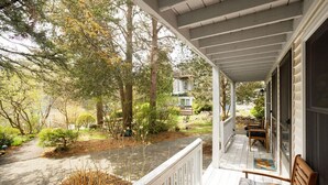 A covered porch with a wooden bench, a pathway leading to a house, and a garden with various plants and trees. - Cozy Nautical Getaway in Cape Neddick (Ogunquit)