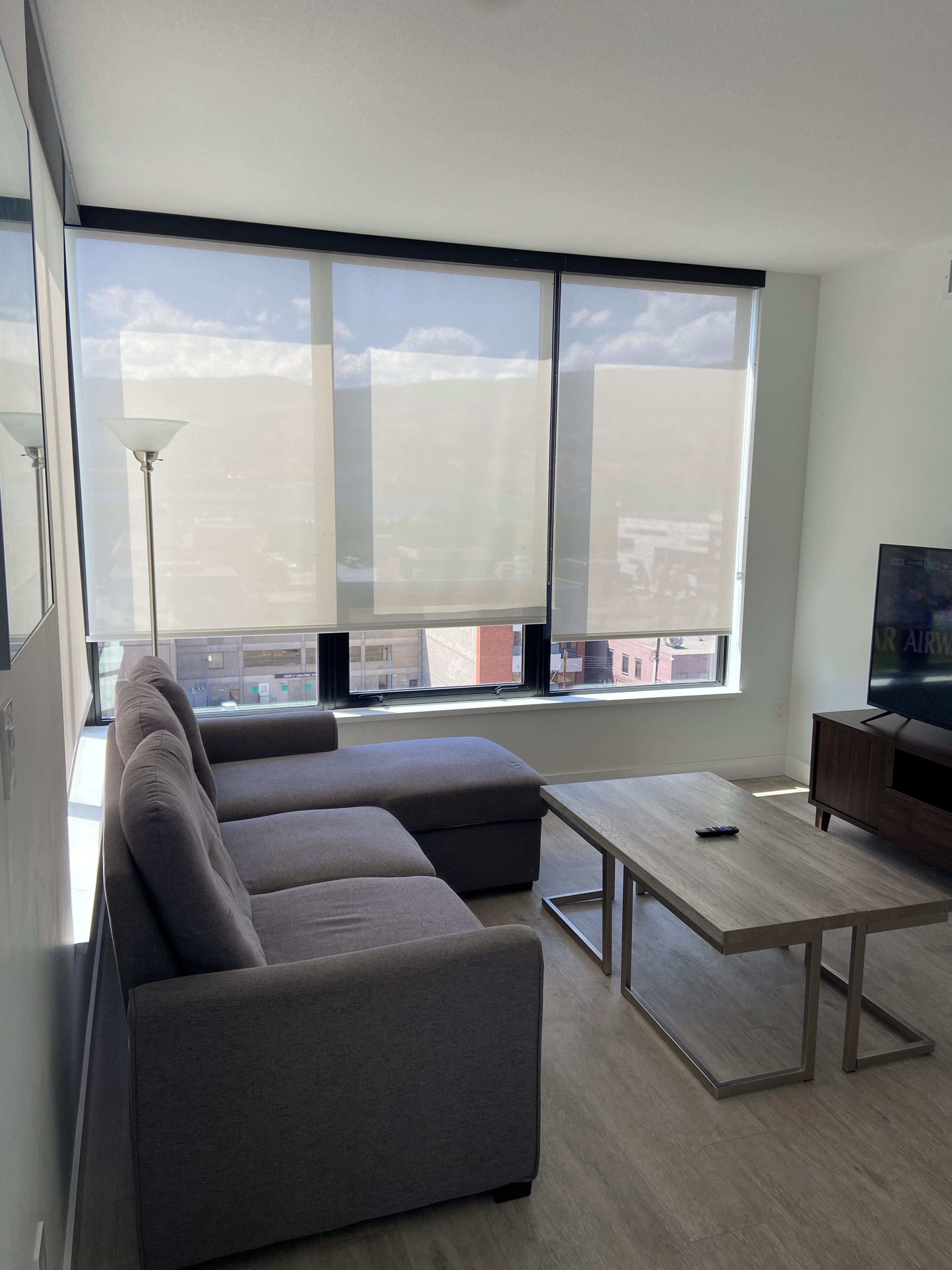 Un salon moderne avec un canapé d’angle, une table basse, un téléviseur posé sur un meuble en bois et de grandes fenêtres offrant une vue sur la ville.