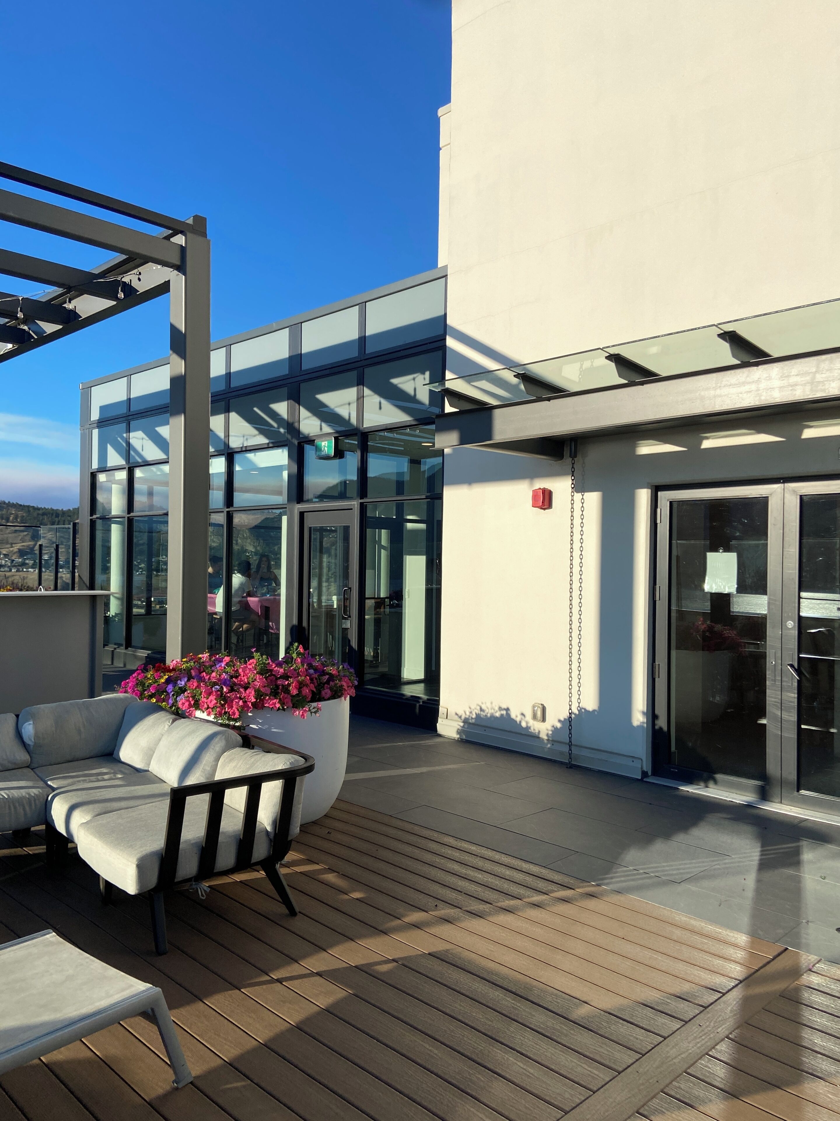 Un bâtiment moderne doté de grandes fenêtres en verre, d’une terrasse en bois et d’un espace extérieur aménagé pour s’asseoir.