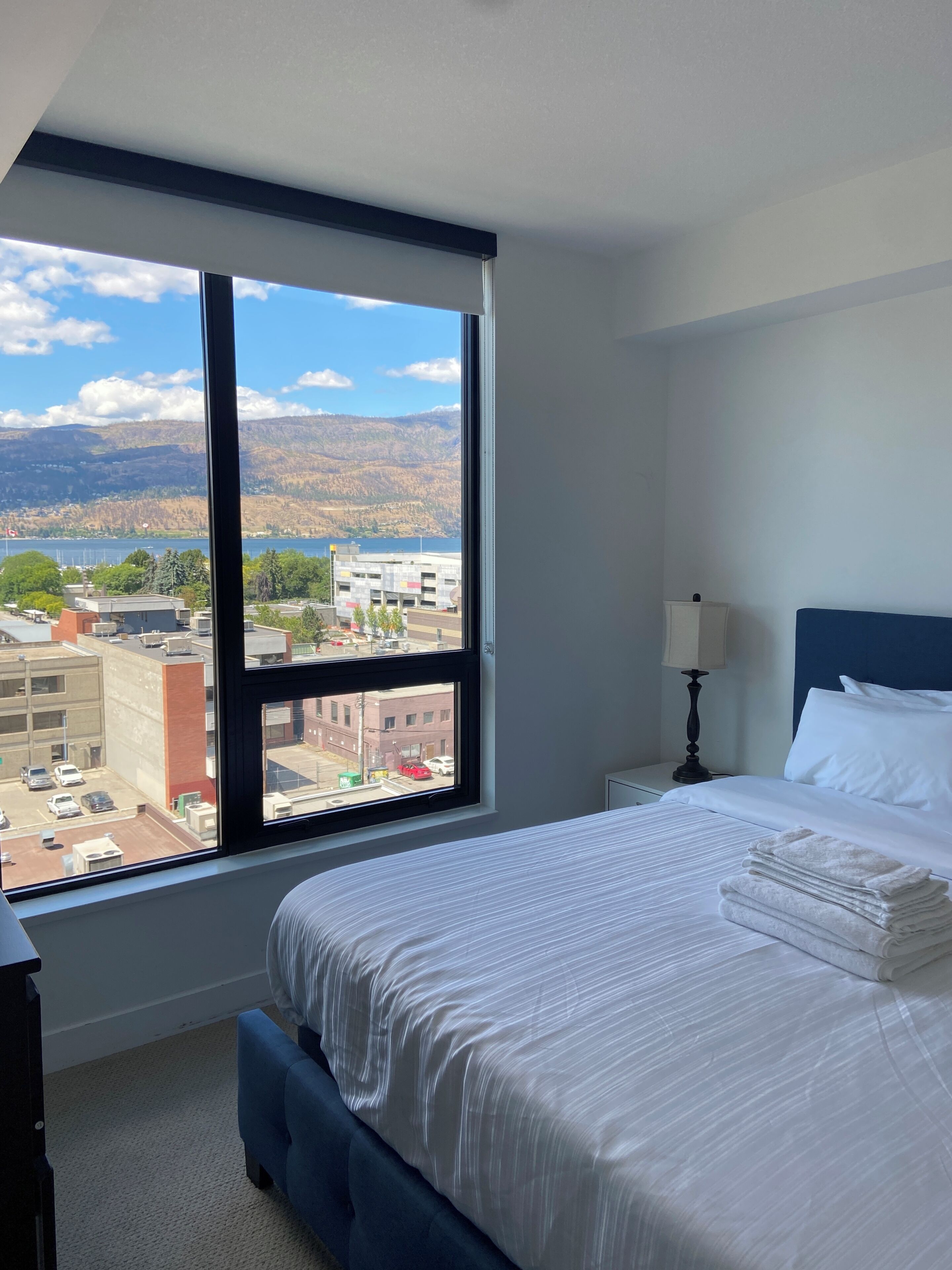 Una habitación de hotel con un ventanal grande, una cama con ropa de cama blanca, cabecera azul y una mesita de noche con lámpara.
