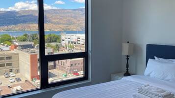 Una habitación de hotel con un ventanal grande, una cama con ropa de cama blanca, cabecera azul y una mesita de noche con lámpara.