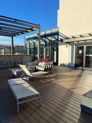 A rooftop terrace with lounge chairs, a seating area, and a glass building with a view.