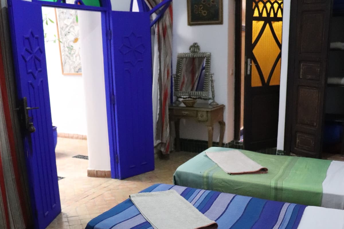 A room with two beds, one with an orange bedspread and the other with a striped bedspread. There is a small table with a round tablecloth in the center of the room.
