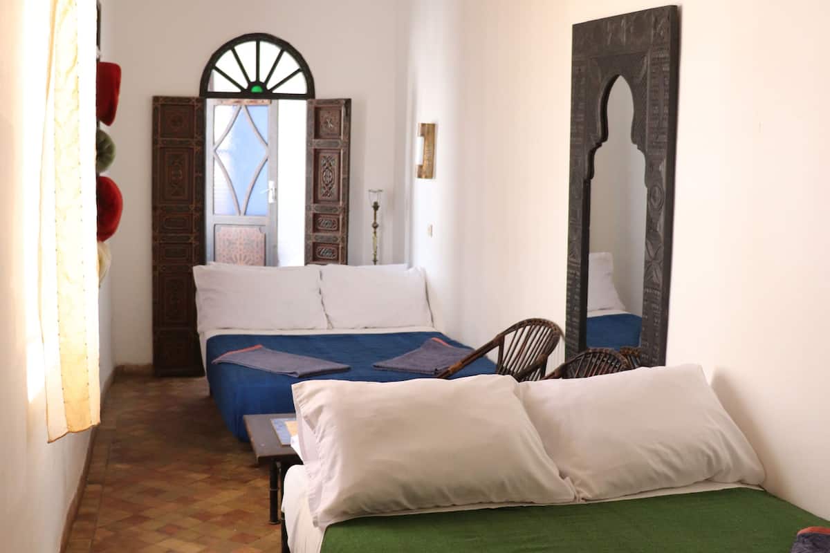A narrow hallway leading to a bedroom with a bed, a chair, a mirror, and a door with intricate designs.