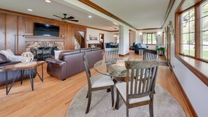A spacious living room with a fireplace, a dining area with a glass table, and a large window offering a view of the outdoors. - Pistakee Lake Estate w/ Boat Dock & Hot Tub! (Johnsburg)