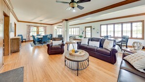 A spacious living room with a wooden floor, a ceiling fan, a dining area with blue chairs, and a large window. - Pistakee Lake Estate w/ Boat Dock & Hot Tub! (Johnsburg)