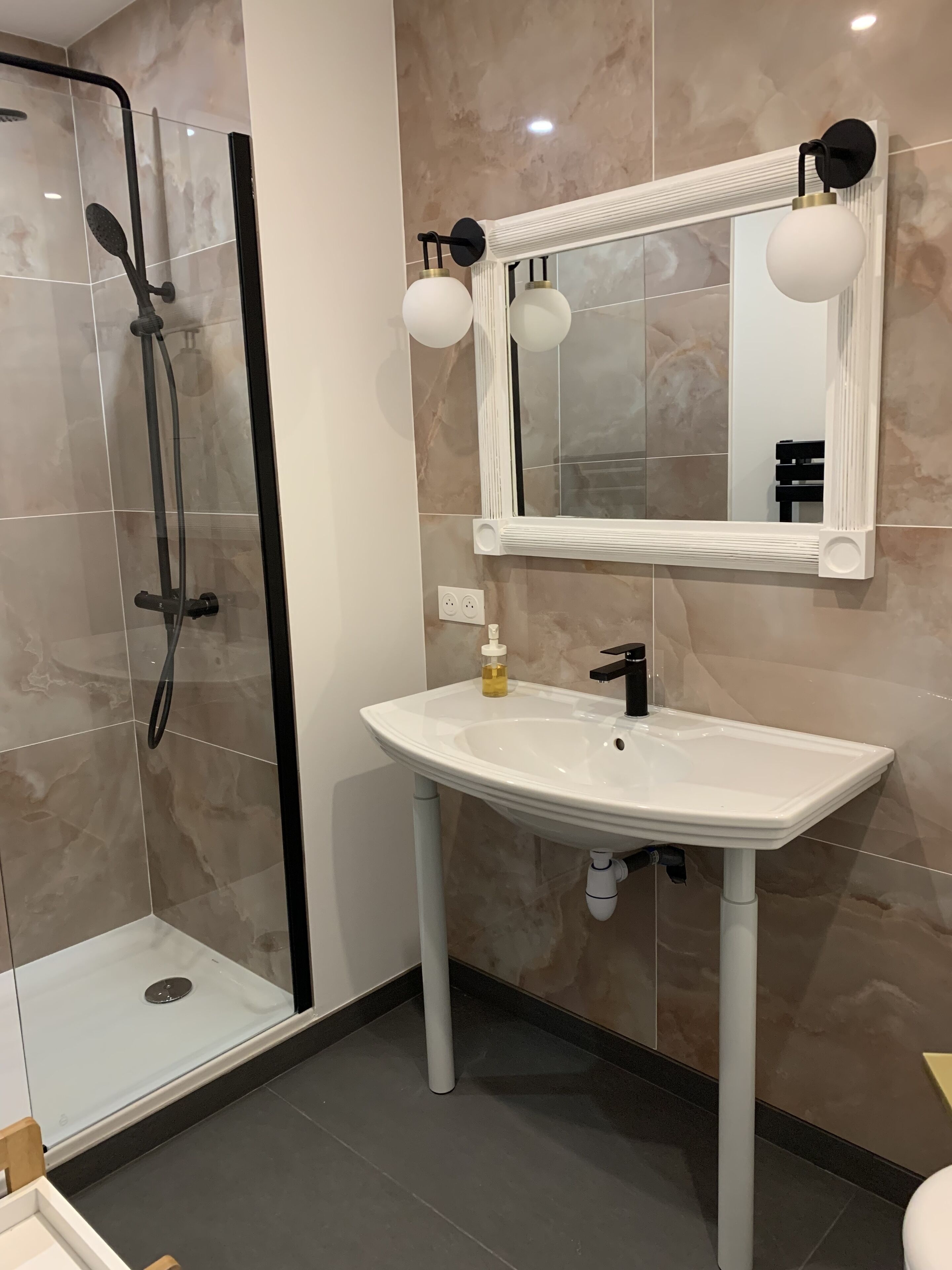 Une salle de bain moderne avec une douche à parois de verre, un lavabo blanc avec un robinet noir et un grand miroir agrémenté de deux appliques murales.