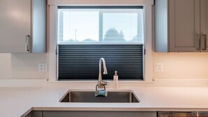 A modern kitchen with a stainless steel sink, a window with blinds, and light-colored cabinets.