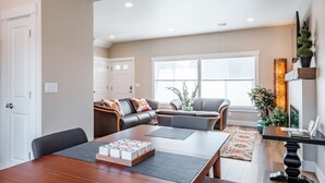 A spacious living area with a wooden dining table, a sofa set, and a large window providing natural light.