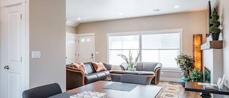 A spacious living area with a wooden dining table, a sofa set, and a large window providing natural light.