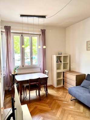 A dining area with a wooden table and chairs, a white cabinet, and a grey sofa. - Devin (Geneva)