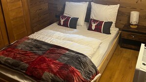 Un dormitorio acogedor con techo de madera, una ventana, una cama con ropa de cama blanca y almohadas rojas y negras, y una mesita de noche con una lámpara.