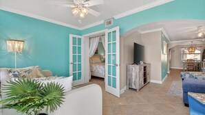 Un vaste espace de vie agrémenté d’un ventilateur de plafond, un coin repas avec une table et des chaises, et une chambre à coucher avec un lit et des rideaux.